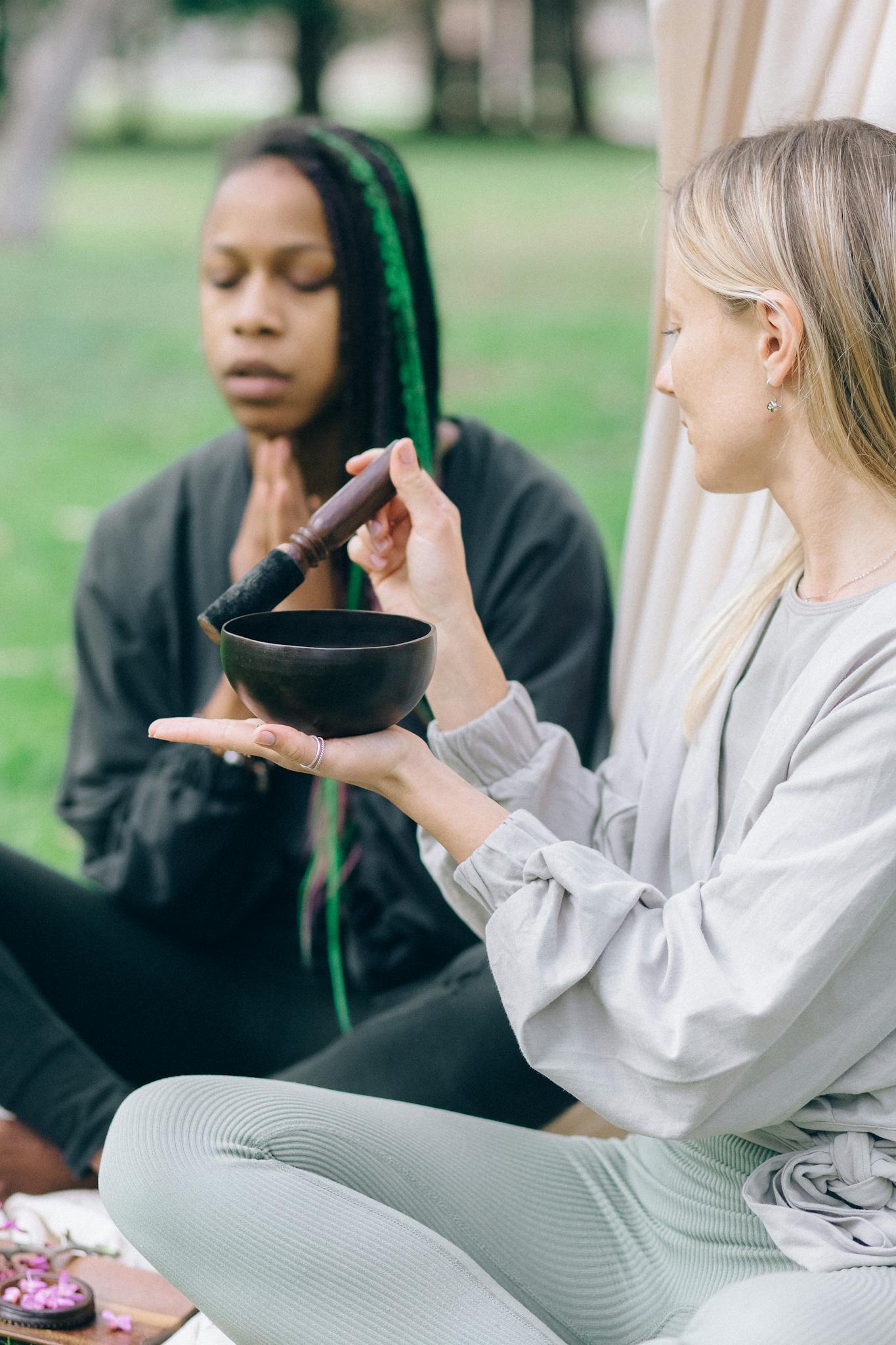 Two women meditate outdoors using a singing bowl, promoting relaxation and wellness.